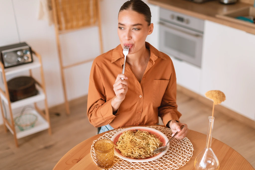 Ava Sinclair thoughtfully tasting a bite of her pasta dish from a fork, with her modern kitchen in the background.
