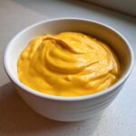 A close-up of rich, bright orange nacho cheese sauce swirled beautifully in a small white bowl.
