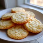 A close-up of several golden-brown sugar cookie cookies piled on a white plate, sprinkled with coarse sugar.