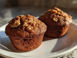 Two freshly baked banana nut muffins topped with walnuts, sitting on a white plate in warm sunlight.