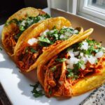 Three golden, slightly crisped chicken tacos filled with shredded chicken and topped with fresh diced white onion and cilantro.