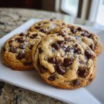 Four freshly baked chocolate chip cookies topped with flaky sea salt on a white plate.