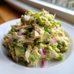 Close-up of a mound of creamy avocado chicken salad mixed with chunks of avocado, red onion, and green onions on a white plate.