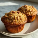 Two golden brown banana nut muffins topped with chopped walnuts sitting on a light plate.