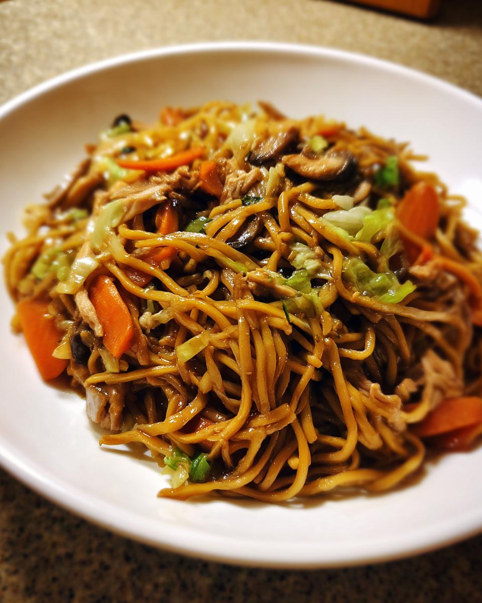 A close-up of a serving of saucy chicken chow mein recipe with noodles, carrots, cabbage, and mushrooms in a white bowl.