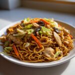 A close-up of a generous serving of chicken chow mein recipe, featuring noodles, sliced chicken, carrots, cabbage, and mushrooms.