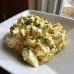 A close-up of chunky, creamy egg salad recipe garnished generously with fresh chopped parsley in a square white bowl.