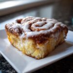 A close-up of a single, square serving of fluffy Cinnamon roll casserole topped with thick white icing.