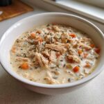 A close-up of a creamy chicken wild rice soup, featuring shredded chicken, carrots, and herbs in a white bowl.
