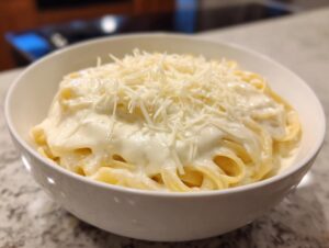 A white bowl filled with fettuccine pasta generously coated in creamy homemade alfredo sauce and topped with shredded Parmesan cheese.