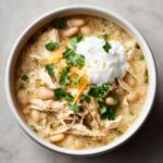 Overhead view of a bowl of creamy crockpot white chicken chili topped with sour cream, shredded cheese, and fresh cilantro.