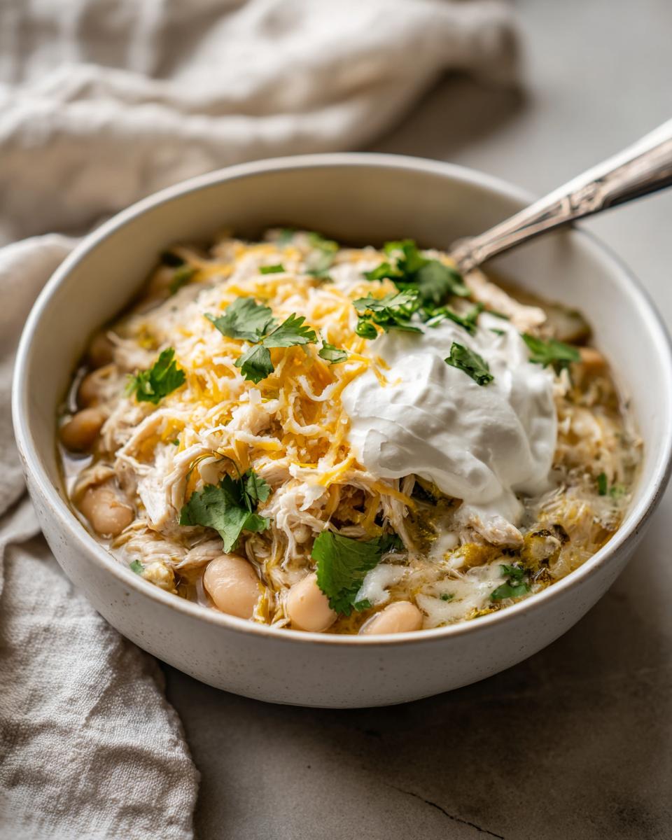 A close-up of a bowl of creamy crockpot white chicken chili topped with shredded cheese, sour cream, and cilantro.