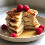 A tall stack of fluffy greek yogurt pancakes, drizzled with syrup and topped with fresh raspberries.