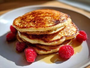 A stack of three fluffy greek yogurt pancakes drizzled with syrup and garnished with fresh raspberries.