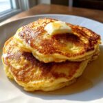Close-up of a stack of golden brown fluffy pancakes topped with melting butter and syrup.