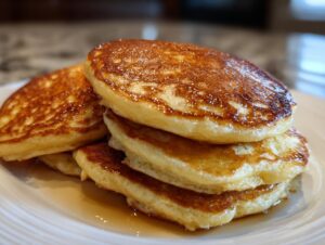 A close-up of a stack of four fluffy sourdough pancakes glistening with syrup on a white plate.