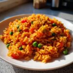 A close-up of fluffy, orange-colored spanish rice mixed with bright green peas and diced red tomatoes on a white plate.