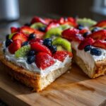 A close-up of a slice of fruit pizza sugar cookie topped with whipped cream, strawberries, blueberries, and kiwi.