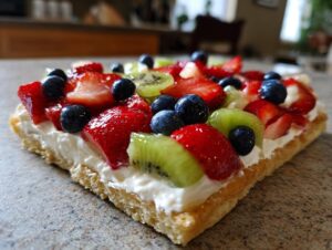 A close-up, low-angle view of a slice of fruit pizza sugar cookie topped with cream cheese frosting and fresh berries.