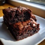 Two fudgy date brownies stacked on a white plate, showing a rich, dark interior and chocolate chips.