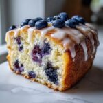 A close-up of a moist slice of blueberry scones loaf, topped with white glaze and fresh blueberries.