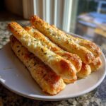 A stack of golden, cheesy garlic parmesan breadsticks sprinkled with parsley on a white plate.