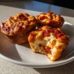 Three golden-brown pizza muffins on a white plate, one of which has a bite taken out, showing the cheesy interior.