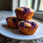 Three golden brown blueberry protein muffins stacked on a white plate, showing juicy berries on top.