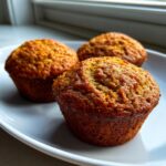 Close-up of three golden brown, moist bran muffins resting on a white plate near a window.
