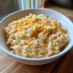 A close-up of a white bowl filled with creamy, comforting cheesy rice topped with melted shredded cheddar.
