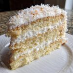 Close-up of a delicious slice of layered coconut cake with white frosting and shredded coconut topping.