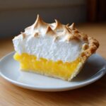 A perfect slice of lemon meringue pie showing bright yellow filling and tall, toasted meringue peaks.