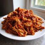 A generous mound of saucy, shredded Slow Cooker Pulled Pork piled high on a white plate near a window.
