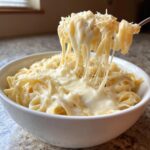A fork lifts fettuccine noodles coated in creamy, stretchy homemade alfredo sauce, topped with grated Parmesan cheese.