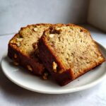 Two moist slices of sugar free banana bread topped with walnuts resting on a white plate.