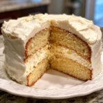 A two-layer vanilla cake recipe with thick white frosting, showing a slice removed to reveal the moist crumb.