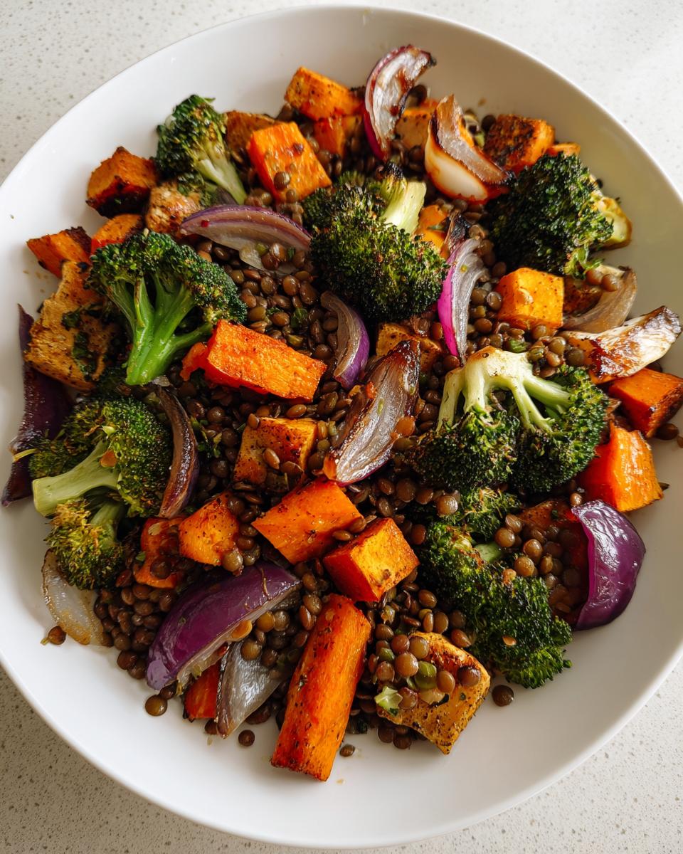 A white bowl filled with a vibrant veggie lentil salad featuring roasted sweet potatoes, broccoli florets, and red onion wedges.