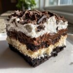 A square slice of rich 4-layer mud pie showing a dark crust, cream layer, chocolate pudding, and whipped topping with chocolate shavings.