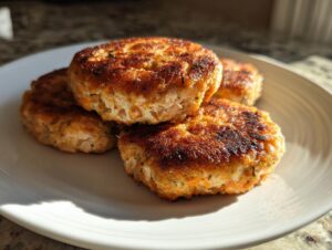 Four perfectly pan-fried, crispy salmon cakes stacked slightly on a white plate, catching bright sunlight.