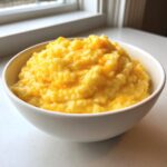A white bowl filled with creamy, bright yellow cheese grits, sitting on a white counter near a window.