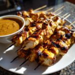 Close-up of grilled chicken satay skewers served on a white plate next to a bowl of peanut dipping sauce.