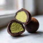 Close-up of amazing dubai chocolate balls, one cut in half revealing a bright green filling inside a dark chocolate shell dusted with cocoa.
