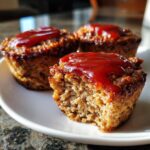 Three individual mini meatloaf portions on a white plate, one bitten into, topped with a glossy red ketchup glaze.