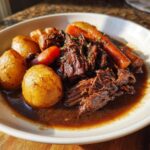 A close-up of tender, shredded pot roast served in rich gravy with whole roasted potatoes and carrots.