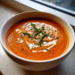 A close-up of a vibrant orange bowl of rich tomato bisque, swirled with cream and topped with fresh basil.