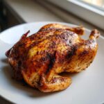 A beautifully roasted whole chicken with dark, seasoned, crispy skin resting on a white plate.