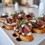 Close-up of several steak crostini appetizers topped with medium-rare steak, white cheese, and rosemary sprigs.