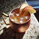 Close-up of a frosty copper mug filled with an apple cider Moscow mule, ice, and garnished with apple slices.