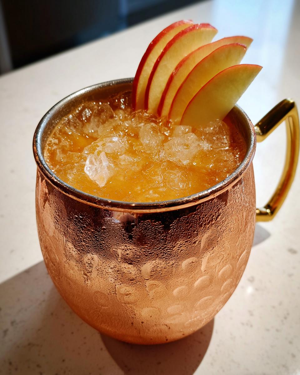 Close-up of a frosty apple cider moscow mule cocktail served in a hammered copper mug, garnished with thin apple slices.