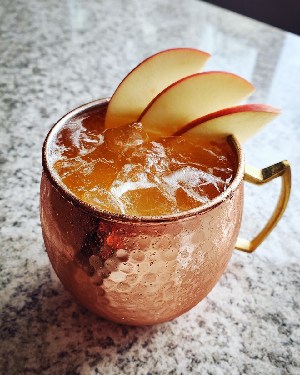 A close-up of an Apple Cider Moscow Mule cocktail filled with ice and garnished with apple slices in a hammered copper mug.
